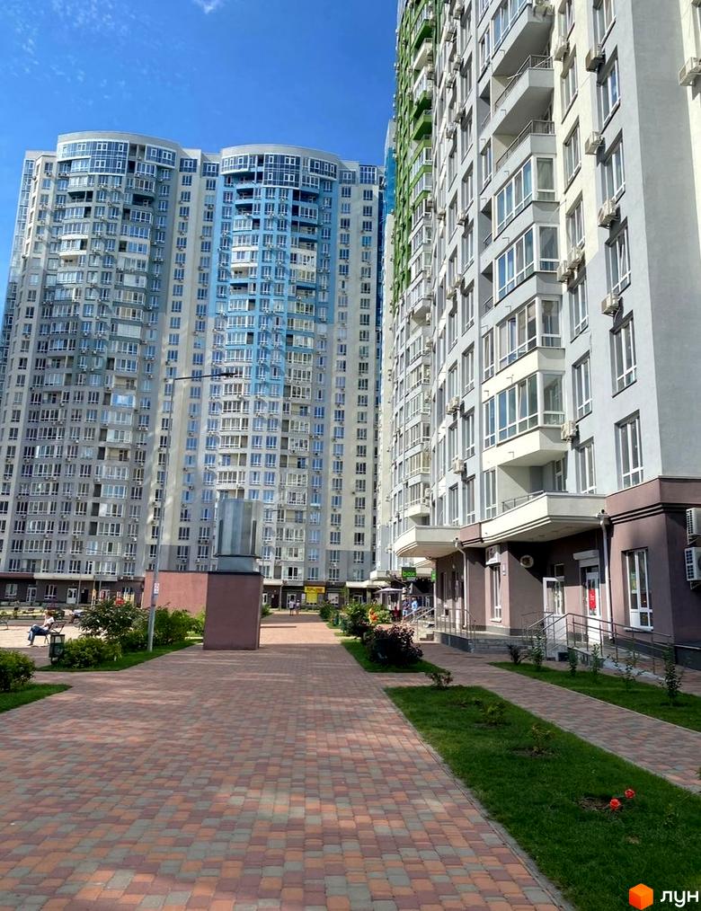 Новобудова ЖК Каховська з блакитними та сірими фасадами, великою кількістю вікон та кондиціонерів. Прибудинкова територія з бруківкою, газоном та кущами.