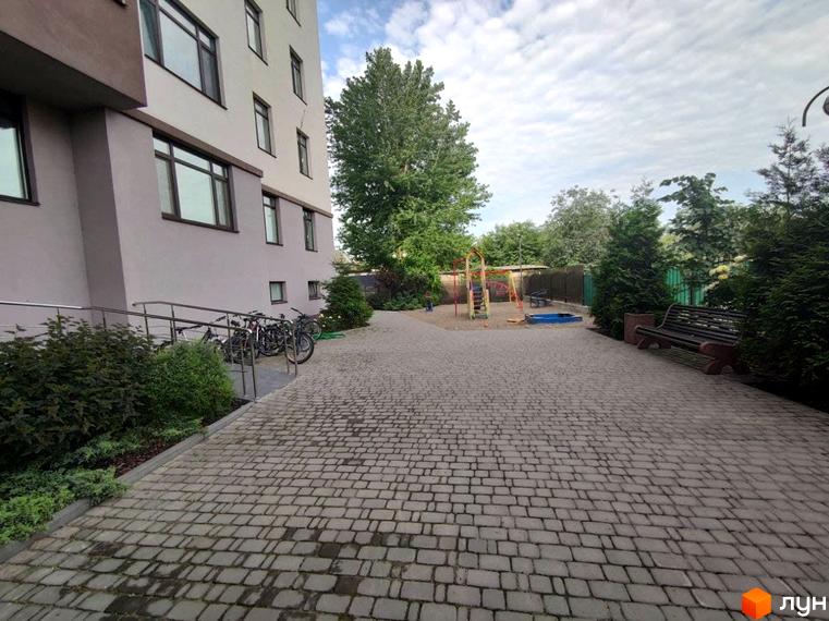 Багатоповерховий будинок ЖБ Light house світлого кольору з темними вікнами та пандусом. Прибудинкова територія з бруківкою, дитячим майданчиком та лавкою.