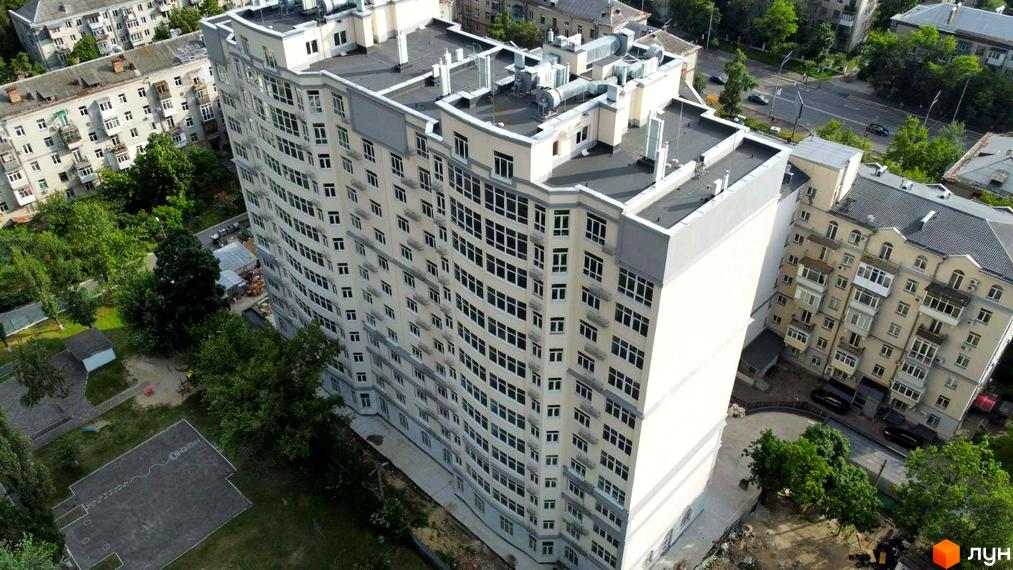 Житловий комплекс Дом на Строителей – сучасна багатоповерхова будівля з вигнутим фасадом та великою кількістю вікон. Навколо будівлі зелена зона з деревами та дитячим майданчиком.