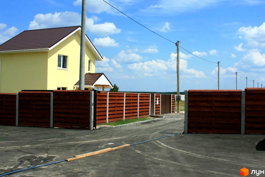 Будинок житлового комплексу КМ Солнечная Поляна з жовтою фасадною частиною, огорожа та асфальтована дорога.