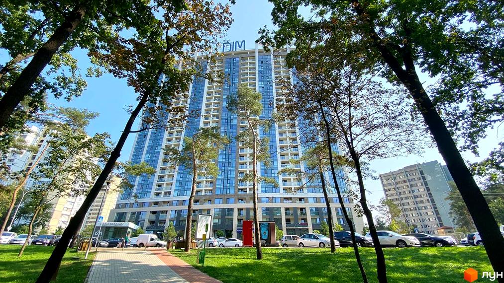 Новобудова ЖК Автограф з блакитними вікнами та балконами, оточена деревами. Прибудинкова територія з доріжкою та автомобілями.