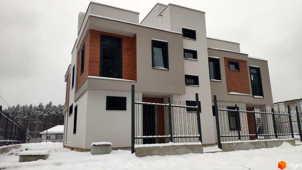 Два дуплекси світлого кольору з темними вікнами та дерев'яними вставками. Територія Дуплексів „Сосновий бір“ засніжена, з металевим парканом та лісом.