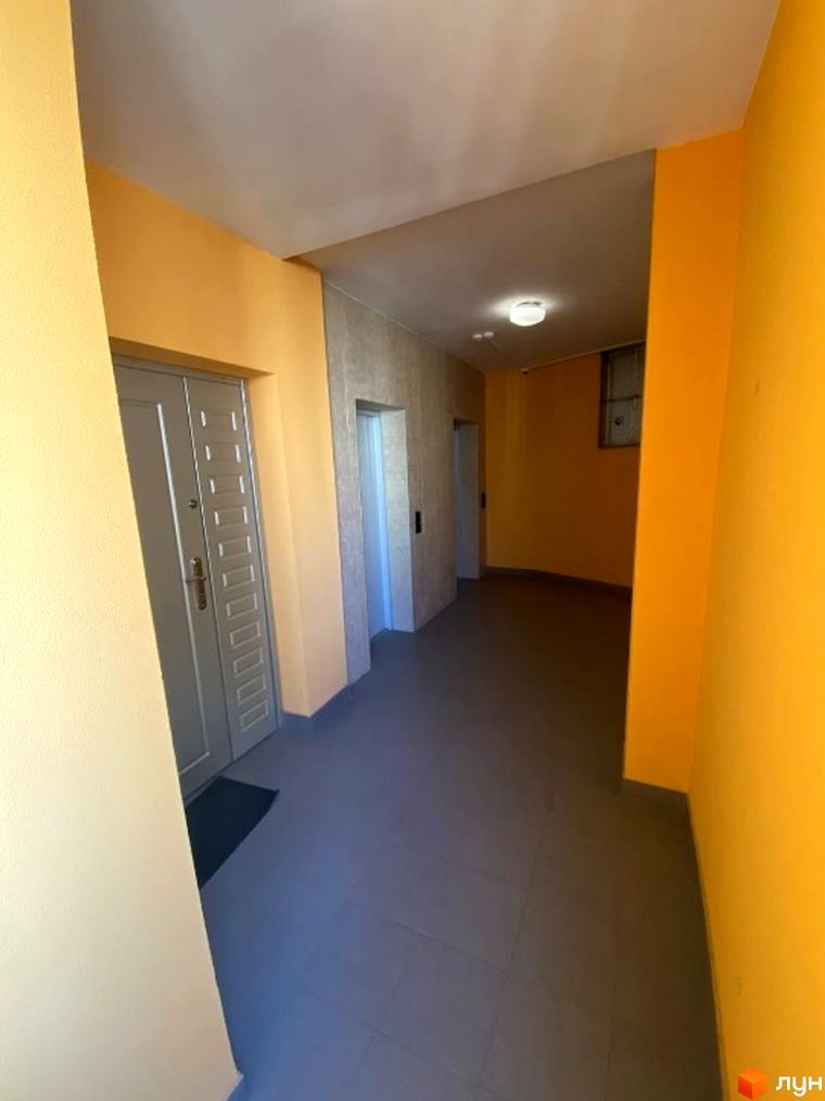 Коридор житлового комплексу з жовтими стінами та плитковою підлогою. Вид на двері квартир та ліфти.