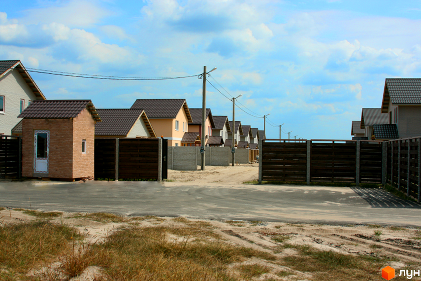 Зображення Котеджного містечка Солнечная Поляна: одноповерхові будинки з коричневими дахами та дерев'яними парканами. На передньому плані асфальтована дорога.