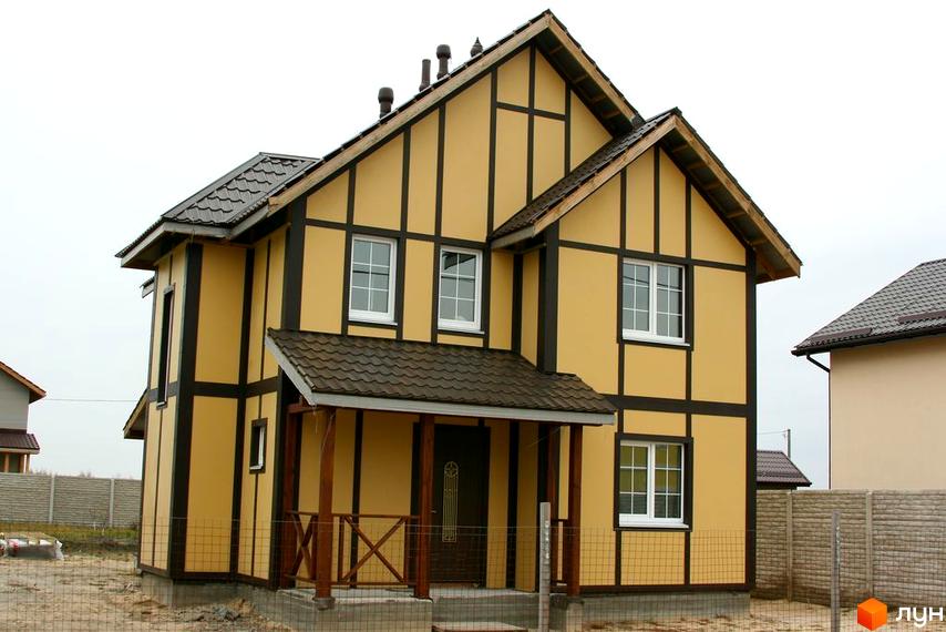 Двоповерховий будинок з жовтими стінами та коричневими балками в Котеджному містечку Солнечная Поляна. Поруч зелена трава та огорожа.