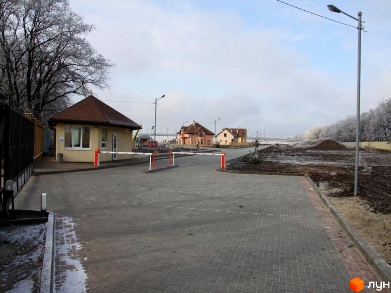 В'їзна зона житлового комплексу КМ Європа з охороною та новими будинками. Прибудинкова територія з асфальтованими доріжками та зеленими насадженнями.