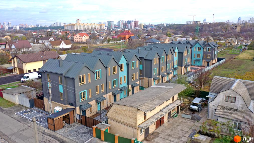 Таунхауси New Smart 13 – це низка двоповерхових будинків з сірими та блакитними фасадами та темними дахами, розташованих вздовж асфальтованої дороги з паркувальними місцями та зеленими зонами.