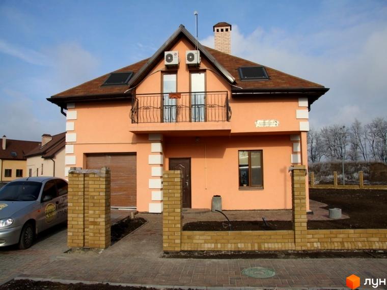 Зображення будинку в Котеджному містечку Європа. Двоповерховий будинок з балконом та коричневим дахом, перед будинком цегляний паркан.