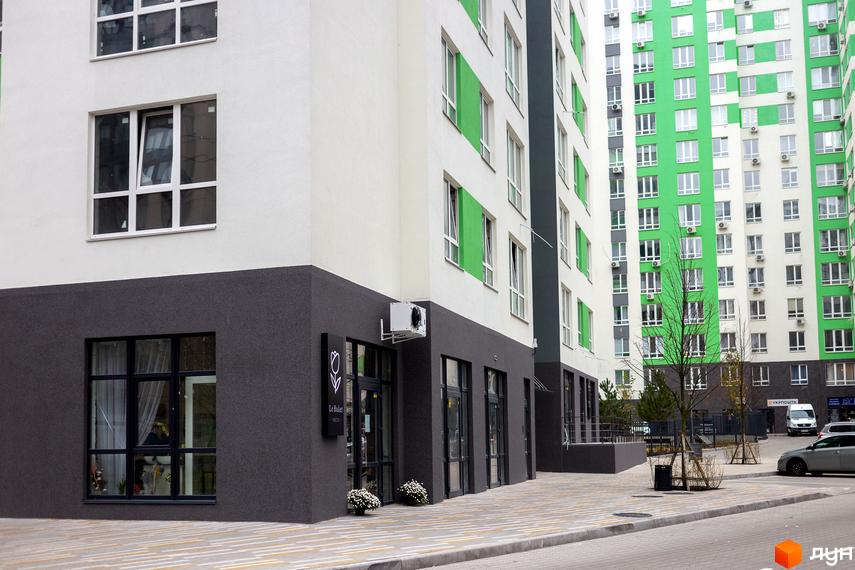 Зовнішній вигляд новобудови ЖК City Park з біло-зеленими фасадами та сірою цокольною частиною. Поруч розташована прибудинкова територія з тротуаром та деревами.