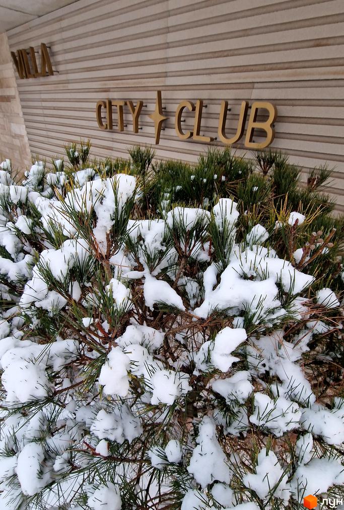 Котеджне містечко Riviera City Park з золотими літерами на фасаді будівлі, оточене засніженими хвойними деревами.