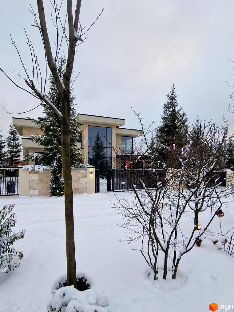 Котеджне містечко Riviera City Park у зимовий період. Фасад будинку з великими вікнами та оздобленням з натурального каменю, навколо засніжена територія з деревами.