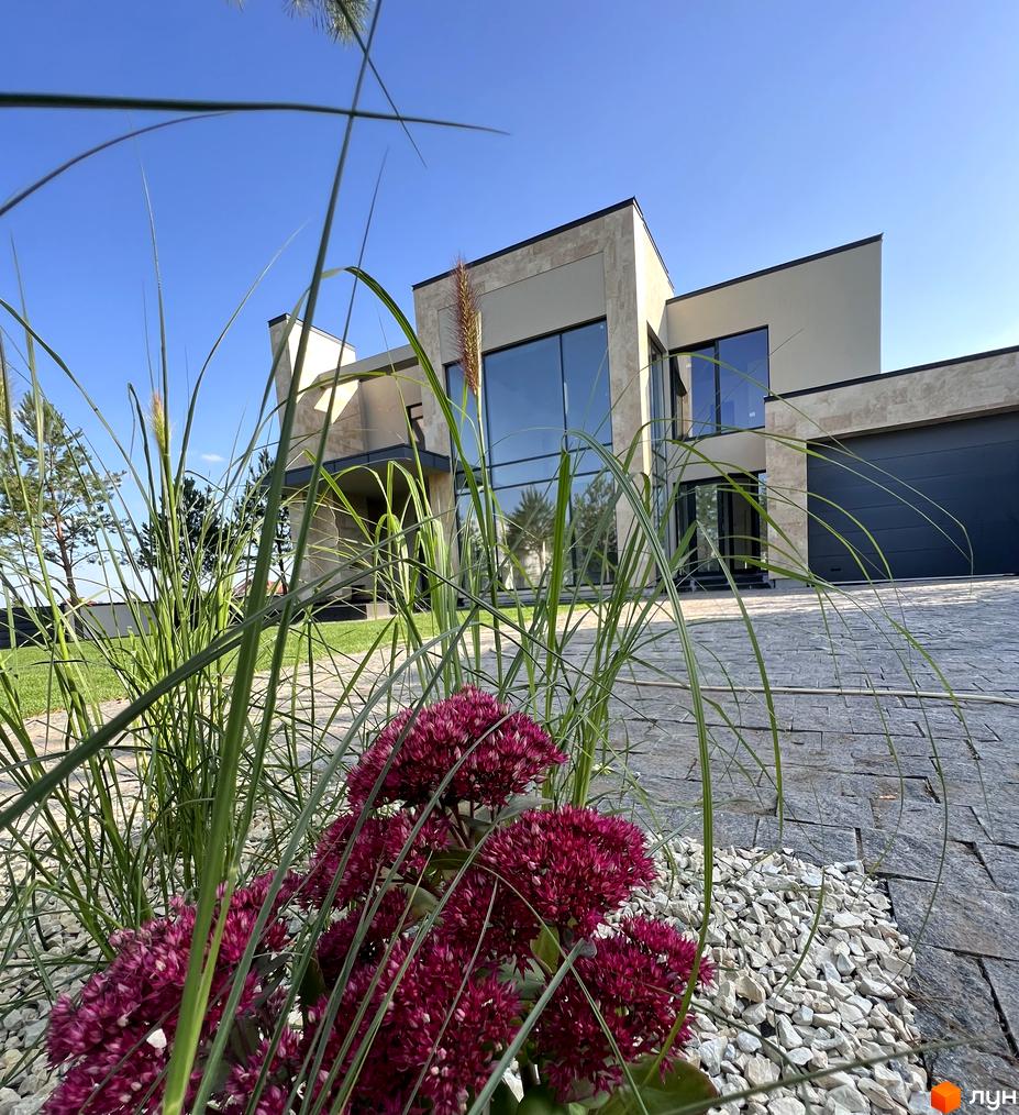 Котеджне містечко Riviera Royal: двоповерховий будинок з великими вікнами та сірою цегляною облицовкою, перед ним зелена галявина та квіти.