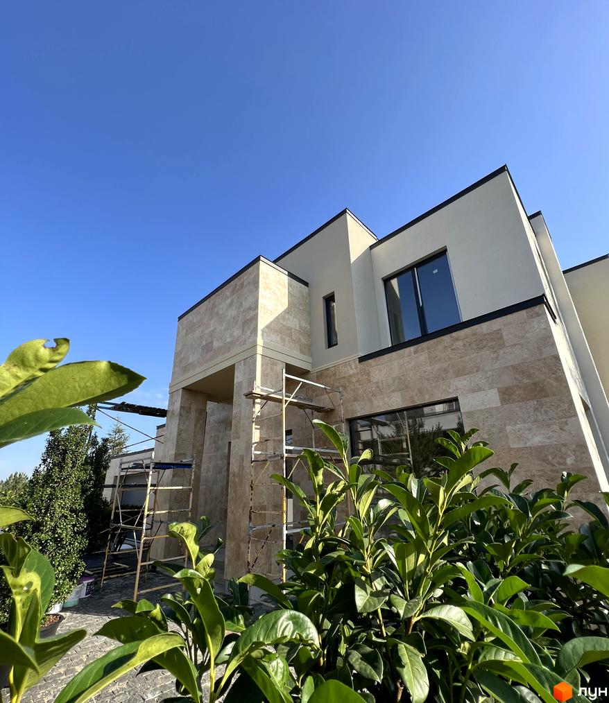 Котеджне містечко КМ Riviera Royal: двоповерховий будинок з бежевим фасадом, оздобленим натуральним каменем, та великими вікнами. Навколо будинку зелені насадження та бруківка.