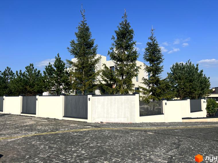 Котеджне містечко Riviera Royal: сучасний будинок з великими вікнами та зеленою територією з хвойними деревами. Навколо будинку розташована огорожа та вимощені доріжки.