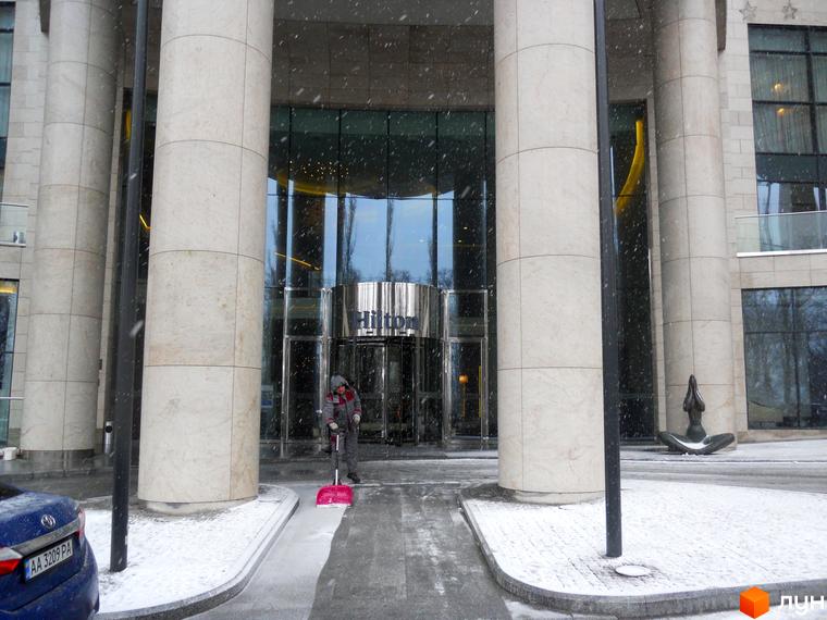 Вхідна група житлового комплексу H-Tower з колонами та скляними фасадами. Прибудинкова територія з засніженим тротуаром.