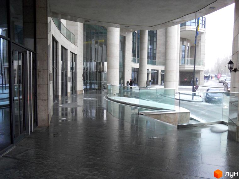 Житловий комплекс H-Tower: архітектурний вигляд будівлі з колонами та скляними фасадами, мокрий тротуар та дорога з автомобілями.