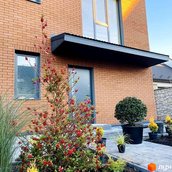 Зовнішній вигляд будинку з цегляним фасадом, вікнами та дверима. На прибудинковій території ростуть рослини з червоними ягодами та зелені кущі.