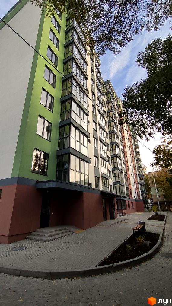 Зовнішній вигляд новобудови на вулиці 600-річчя з яскравим фасадом та скляними балконами. Прибудинкова територія з лавкою та зеленими насадженнями.