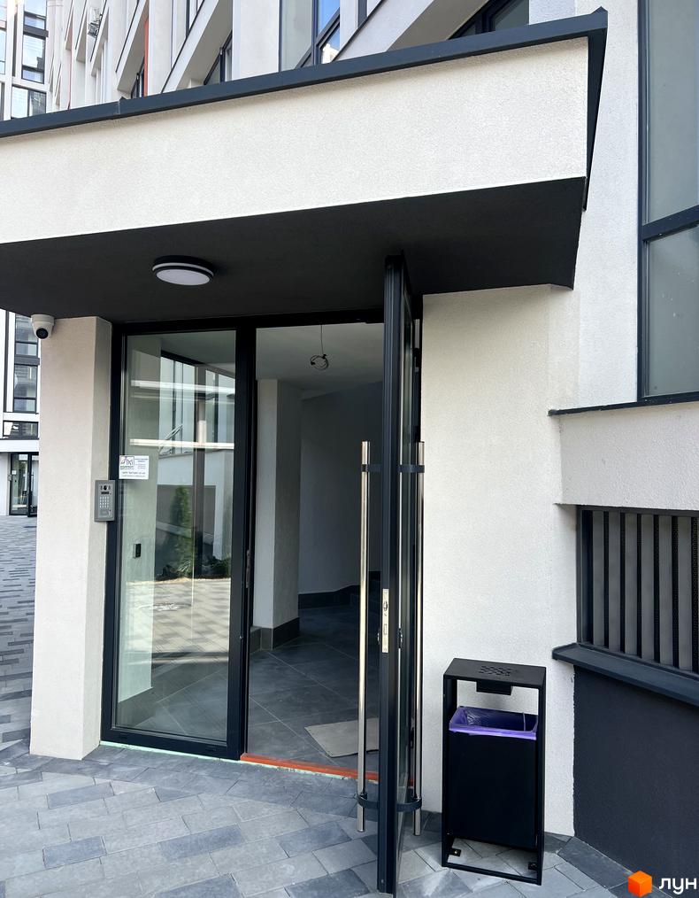 Вхід до Житлового комплексу City Line з білим фасадом, темним навісом та прозорими скляними дверима. Перед входом сіра бруківка.