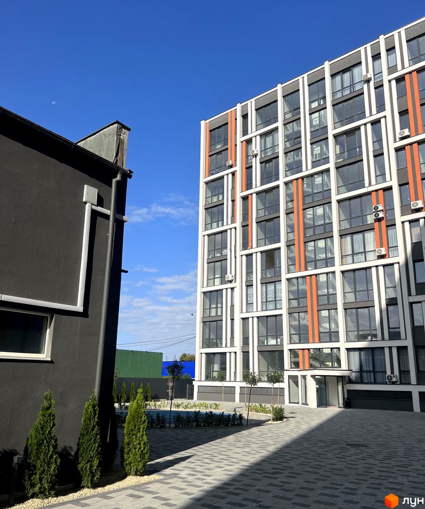 Фасад ЖК City Line з великими вікнами, білими рамами та помаранчевими вертикальними смугами. На подвір'ї укладена бруківка та висаджені дерева.