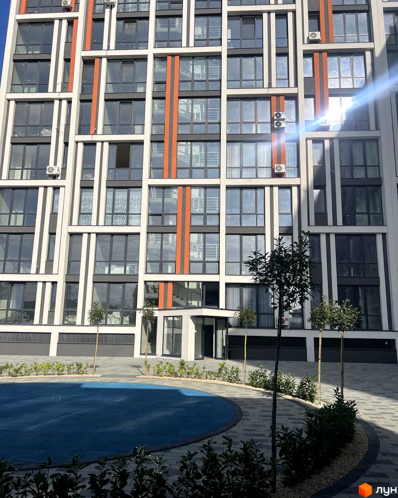 Фасад Житлового комплексу City Line має сіро-білі та помаранчеві вертикальні елементи. На прибудинковій території синій майданчик та доріжки з озелененням.