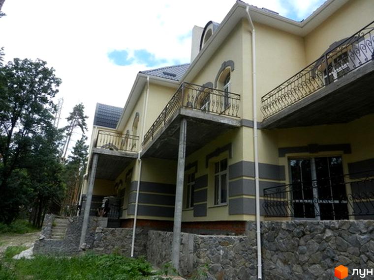 Зображення Котеджного містечка Сосновий бір: двоповерховий будинок з балконами та кам'яним фундаментом. Навколо зелені дерева та доглянута територія.