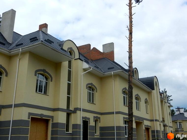 Зовнішній вигляд Котеджного містечка Сосновий бір: двоповерхові будинки з сірими дахами та світло-жовтими стінами. Біля будинків росте високе дерево.