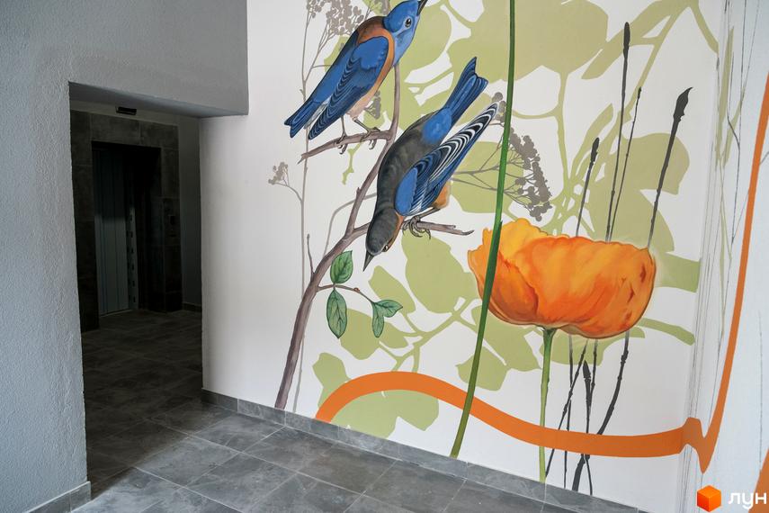 Вхідний хол Житлового комплексу Сонячна Долина з ліфтом та яскравим настінним малюнком птахів і квітів.