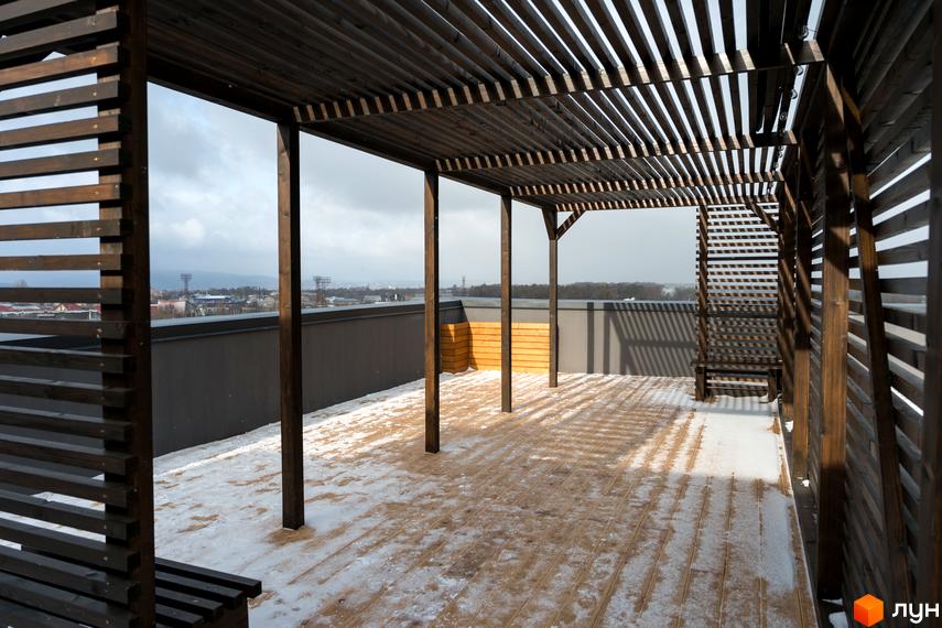 Вид на терасу на даху Житлового комплексу Сонячна Долина з дерев'яною перголою та підлогою, частково вкритою снігом, на тлі міського пейзажу.