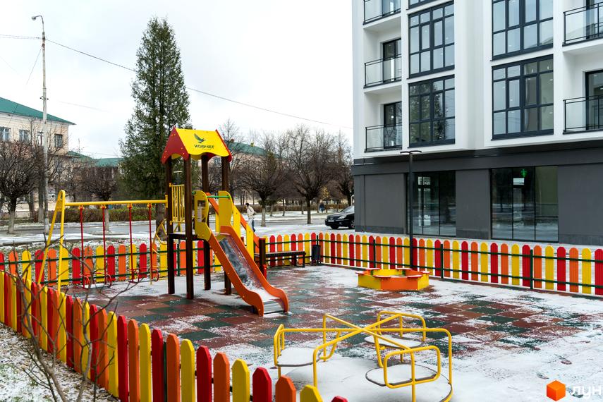 Житловий комплекс Сонячна Долина: багатоповерховий будинок з білим фасадом та великими вікнами. На передньому плані дитячий майданчик з яскравим обладнанням та огорожею.