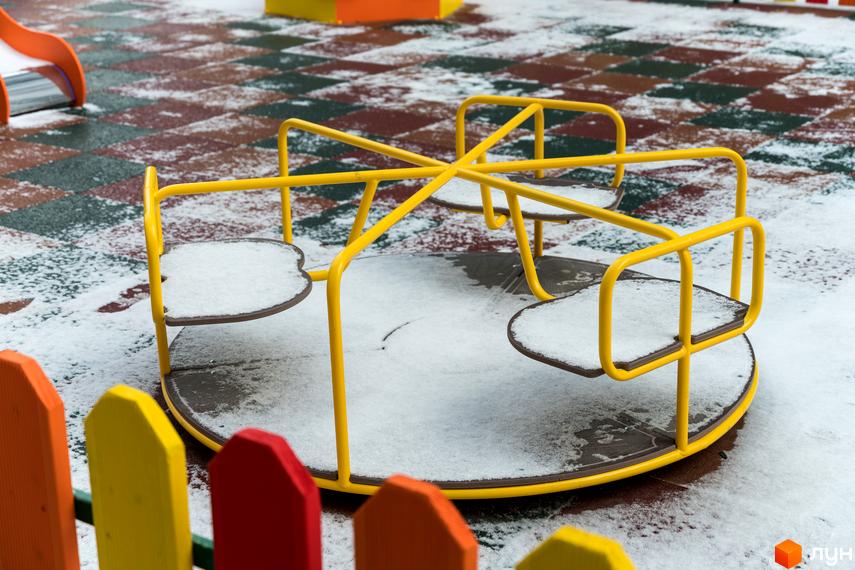 Дитячий майданчик з жовтою каруселлю та різнокольоровим парканом, присипаний снігом, на території Житлового комплексу Сонячна Долина.