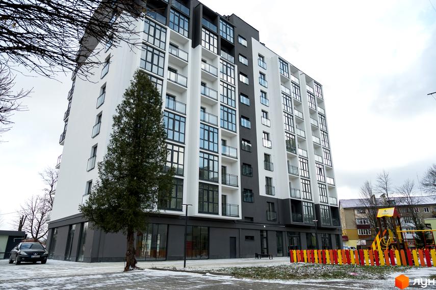 Багатоповерховий будинок Житлового комплексу Сонячна Долина у білих та темно-сірих тонах з балконами. Поруч дитячий майданчик та дерево.