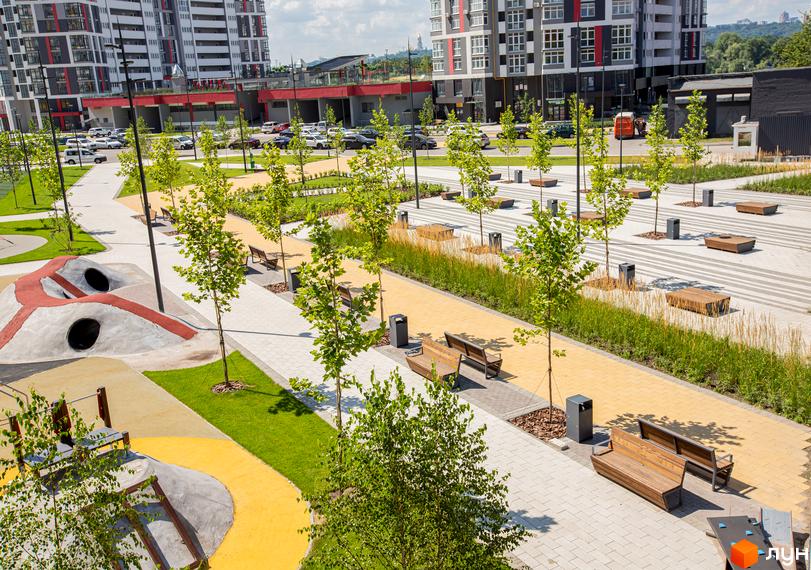 Житловий комплекс Русанівська Гавань. Високі будинки з фасадами сірого та червоного кольорів. На території облаштовані зони відпочинку з лавками, деревами та дитячим майданчиком.