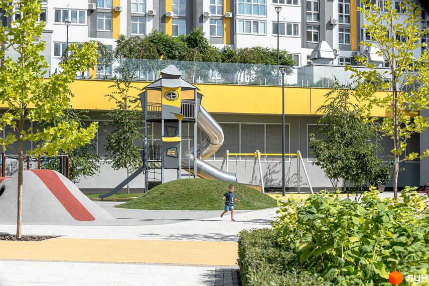 Дитячий майданчик ЖК Русанівська Гавань з гіркою та гойдалками на фоні житлового будинку. На території комплексу висаджені дерева та кущі, а також є пішохідні доріжки.