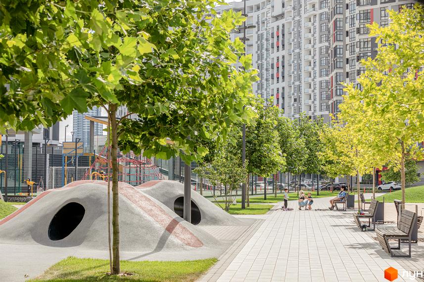 Багатоповерховий житловий будинок Русанівська Гавань з біло-сірим фасадом. Прибудинкова територія облаштована зеленими насадженнями, дитячим майданчиком та доріжками.