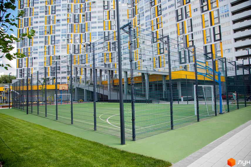 Багатоповерховий будинок Житлового комплексу Русанівська Гавань з біло-сірим фасадом та жовтими вертикальними вставками. Поруч розташований спортивний майданчик із зеленим покриттям та огорожею.