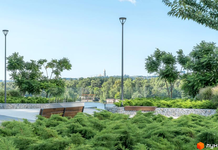 На фото зображено частину житлового комплексу Русанівська Гавань. Видно лавки, ліхтарі та зелені насадження на території біля води.