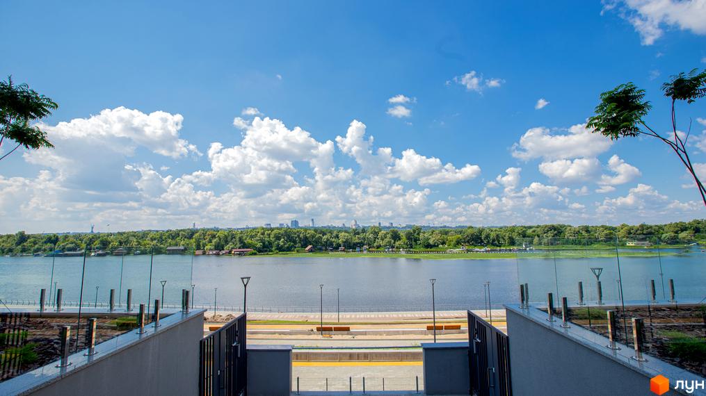 Вид з території Русанівська Гавань на річку та міський пейзаж. На передньому плані видно набережну зі скляними огорожами та лавками.