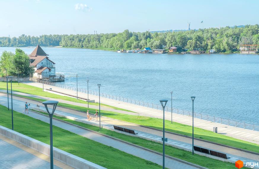 Будівля з коричневим дахом та великими вікнами на березі річки, поруч з упорядкованою набережною Русанівська Гавань.