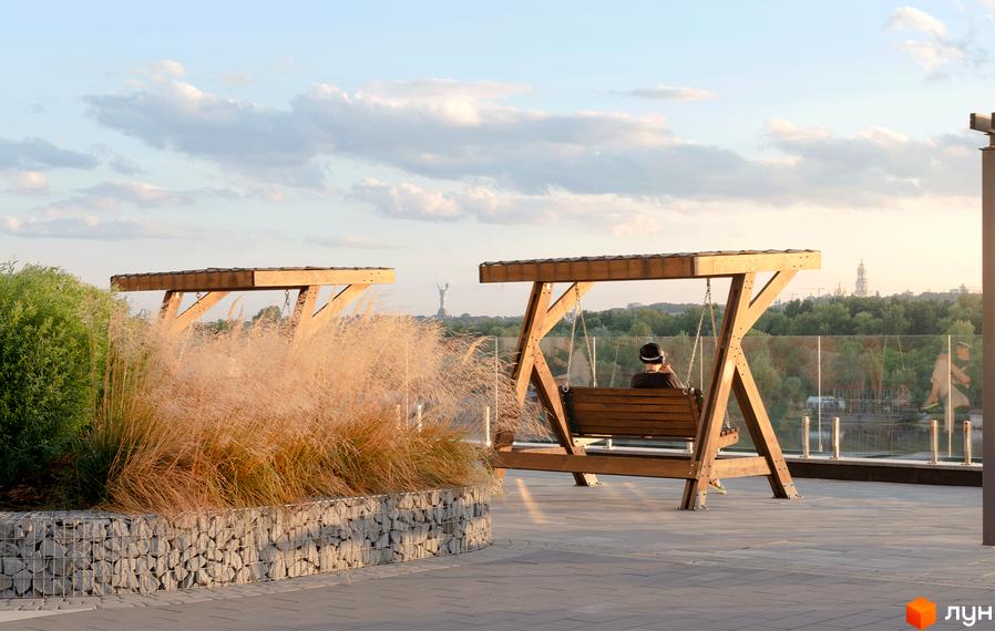 Зона відпочинку житлового комплексу Русанівська Гавань з дерев'яними гойдалками та озелененням. Відкривається вид на річку та міські пейзажі.
