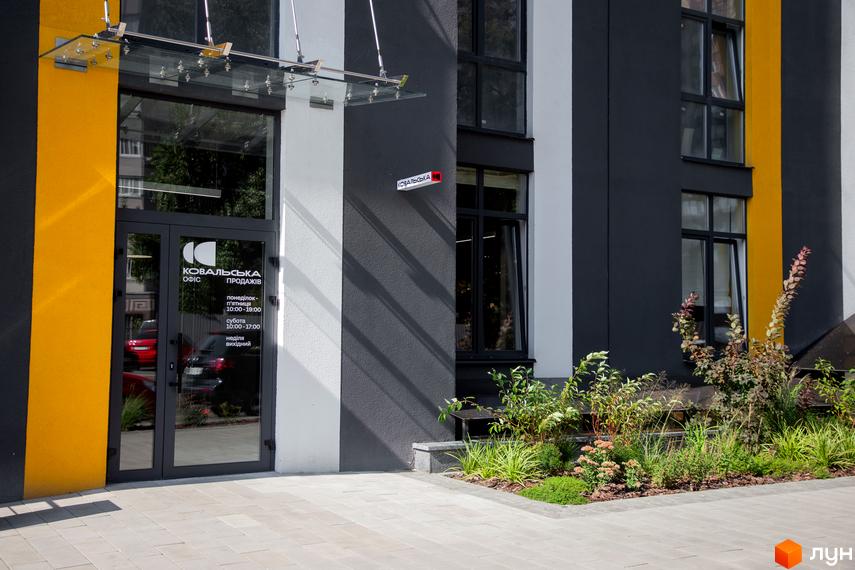 Вхід до офісу продажів житлового комплексу. На фасаді будівлі вивіска з назвою та графіком роботи. Поряд з входом облаштована невелика клумба з рослинами.