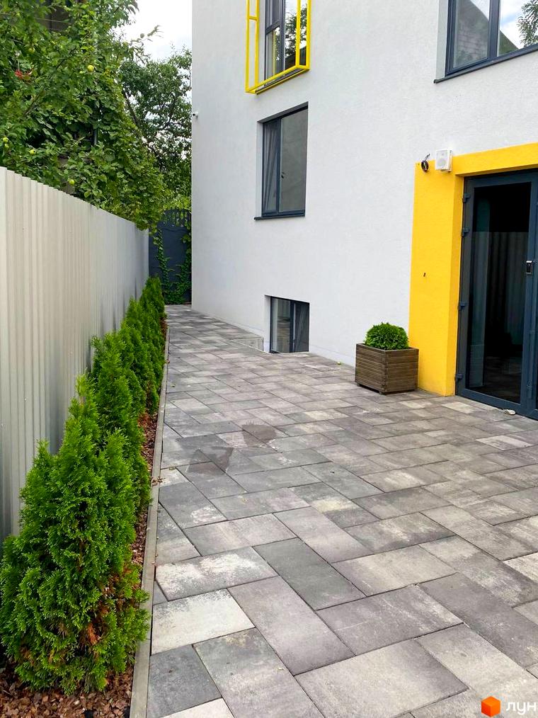 Зовнішній вигляд будинку Coliving ONE Nivki з білими стінами та жовтими елементами. Поряд розташована вимощена доріжка та зелені туї.