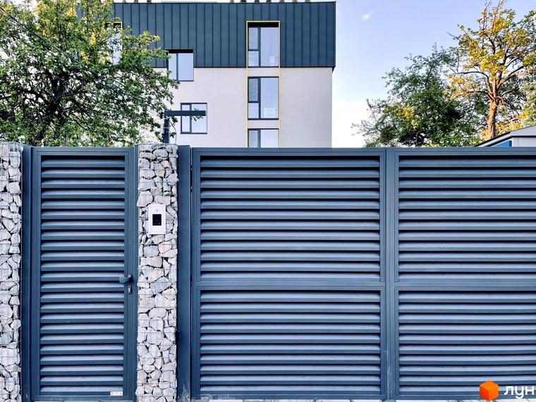 Coliving ONE Nivki: будинок зі світлим фасадом і темним дахом на тлі дерев. Сірі ворота з кам'яними опорами.