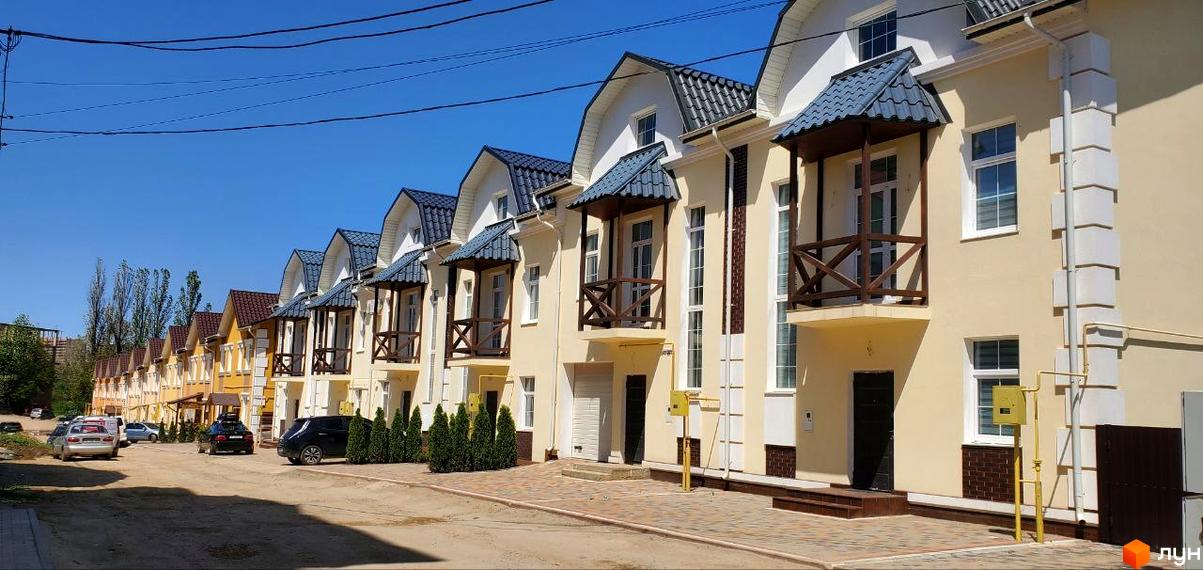 Ряд таунхаусів Гарден Хаус з бежевими фасадами, синіми дахами та дерев'яними балконами. Перед будинками розташована дорога та зелені насадження.