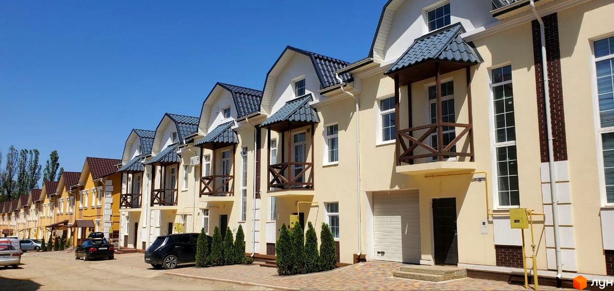 Таунхаус „Гарден Хаус'': ряд будинків світло-жовтого кольору з темно-сірими дахами та дерев'яними балконами. Прибудинкова територія з зеленими насадженнями та проїжджою частиною.