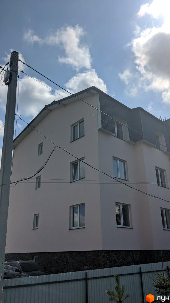 Зовнішній вигляд триповерхової новобудови з великою кількістю вікон та темним дахом. Навколо будинку видно паркан та дерева.