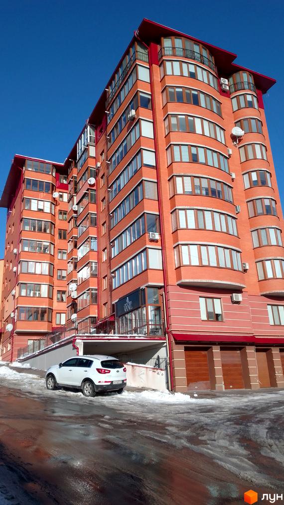 Зовнішній вигляд цегляного житлового комплексу з червоним дахом та численними вікнами, поруч білий автомобіль на засніженій дорозі.