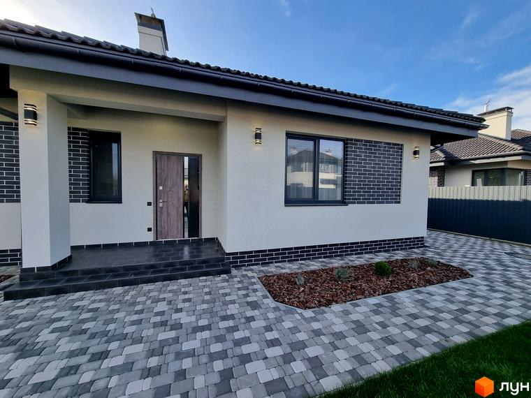 КМ Сонячний: фасад будинку зі світлою штукатуркою, темною цегляною обробкою та вхідними дверима. Прибудинкова територія з бруківкою та клумбою.