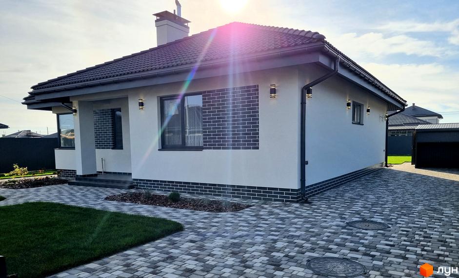 Котеджне містечко КМ Сонячний. Зовнішній вигляд будинку з білою штукатуркою, сірим дахом та декоративним оздобленням фасаду. Прибудинкова територія з бруківкою та зеленим газоном.