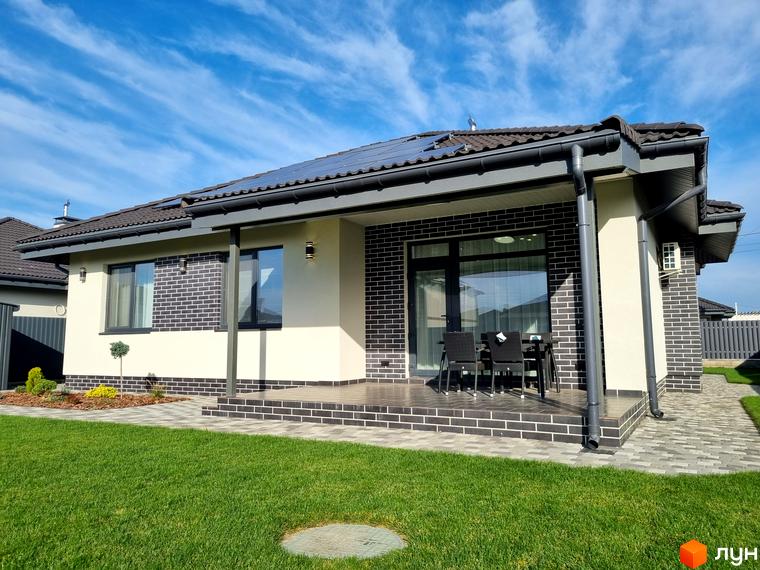 Котеджне містечко КМ Сонячний: одноповерховий будинок з темною цегляною обробкою, великими вікнами та сонячними панелями на даху. Навколо будинку доглянутий газон та бруківка.